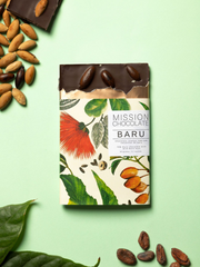 Embalagem do chocolate de BARU com folhas e a castanha sobre um fundo verde com um pouco da barra fora da embalagem, ao redor tem cacau, duas folhas e a castanha.